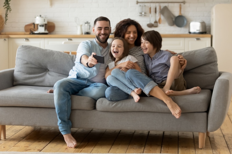Smiling parents with little kids laughing using smartphone together sitting on couch at home.